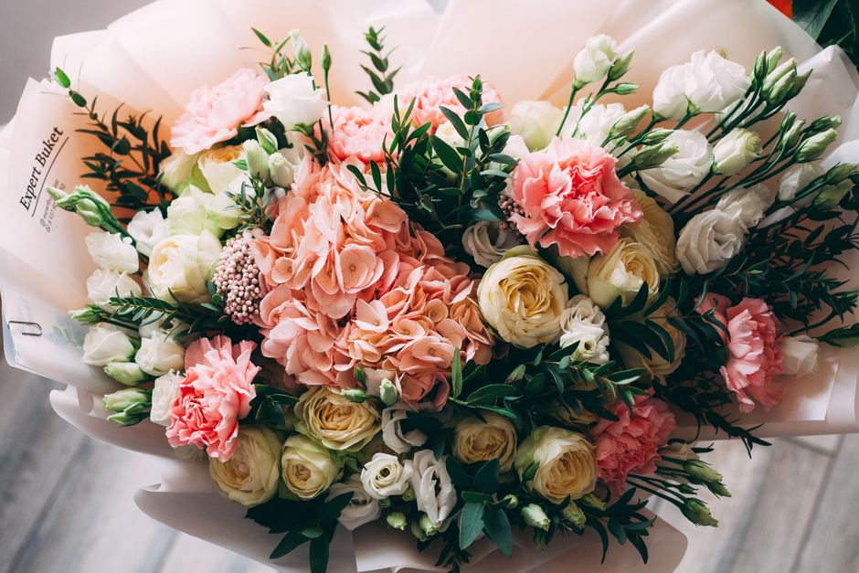 A beautifully arranged floral bouquet featuring soft pink hydrangeas, creamy white roses, and blush pink carnations, complemented by sprigs of greenery and small white filler flowers. The bouquet is w