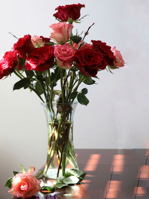 A clear glass vase filled with a mixed bouquet of fresh roses in shades of deep red, blush pink, and soft peach, arranged with green stems and leaves. The flowers are positioned on a wooden surface ne