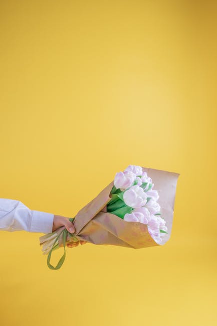 A person, dressed in a white shirt, is holding a bouquet of pale pink tulips wrapped in light brown kraft paper with a green ribbon against a bright yellow background. The tulips have smooth, slightly