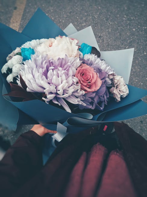 A hand holding a bouquet of fresh flowers wrapped in blue and white paper outside on a paved surface. The bouquet features a large white chrysanthemum, soft pink rose, purple lisianthus, and smaller w