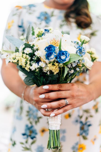 A person holding a freshly crafted bouquets of flowers, featuring white roses, blue roses, small white daisies, and yellow accents, arranged with lush green foliage. The bouquet is wrapped with elegan