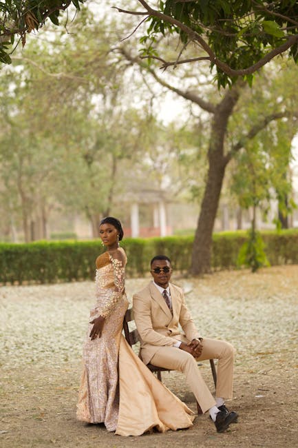 A young woman and a man are seated on a park bench beneath a large leafy tree in an outdoor setting with a soft, natural light. The woman is dressed in an elegant, off-the-shoulder gown with intricate